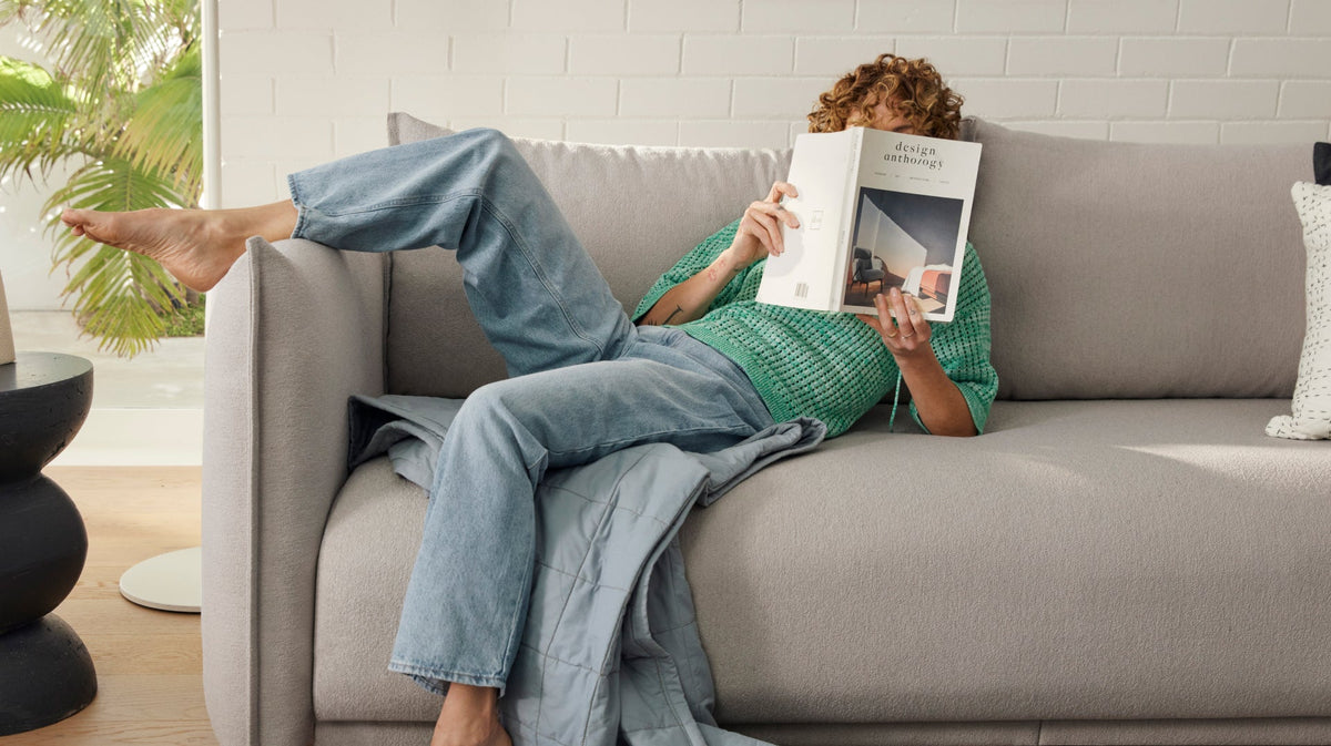 A person with curly hair reads a magazine while lounging barefoot on a light grey sofa, draped with a soft light blue blanket.