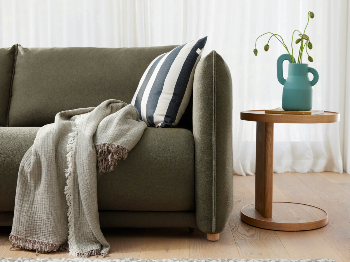 An olive green sofa with a striped black and white pillow and a light grey blanket. A round wood side table holds a teal vase with greenery.