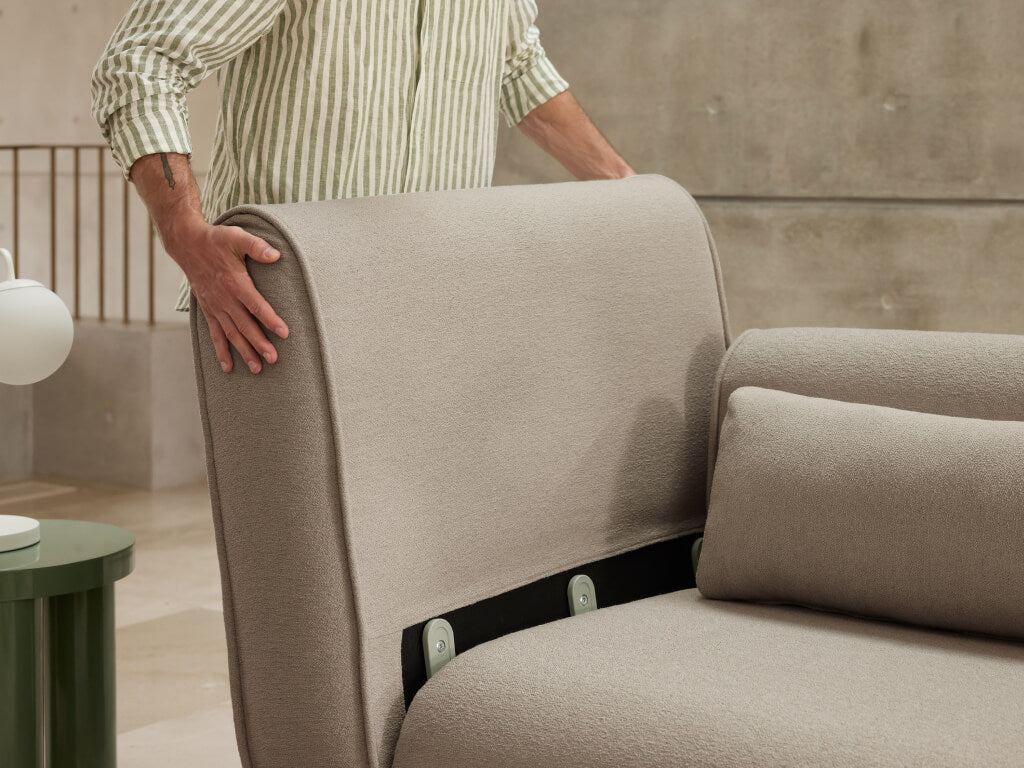 Modern beige upholstered sofa with detachable backrest, featuring plush cushions. Positioned in a minimalist room with stone walls and a green side table.