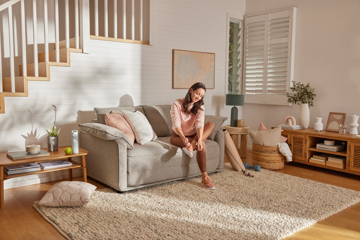 A woman in a light pink sweatshirt and brown leggings sits on a light grey sofa, tying white sneakers in a bright, airy room.