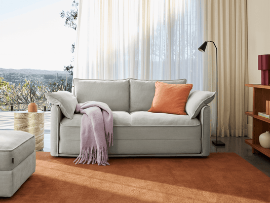 Luxurious light grey sofa in soft fabric, featuring plush cushions and clean lines. Positioned in a modern living room with sheer curtains and a scenic view.