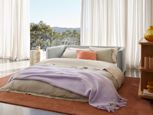 Luxurious grey fabric daybed with plush cushions, set in a bright room featuring floor-to-ceiling windows and scenic views. Paired with stylish side table.