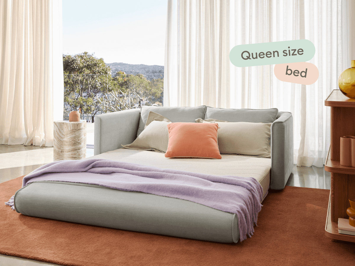 Modern queen-size sofa bed in soft grey fabric, accented with plush cushions. Set on a rich rust rug; elegantly framed by sheer curtains and natural light.