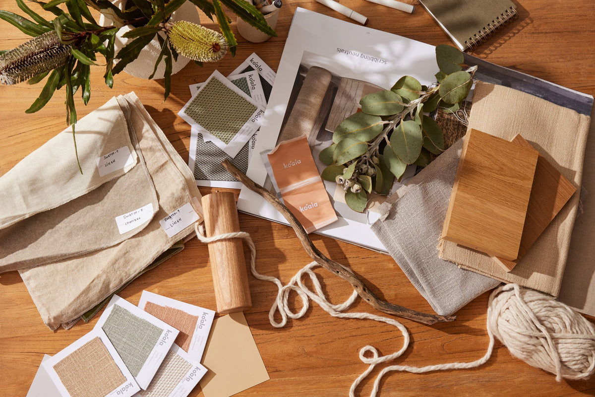 A wooden table displays beige linen fabric, wood samples, greenery, and color swatches on white paper in soft, natural lighting.