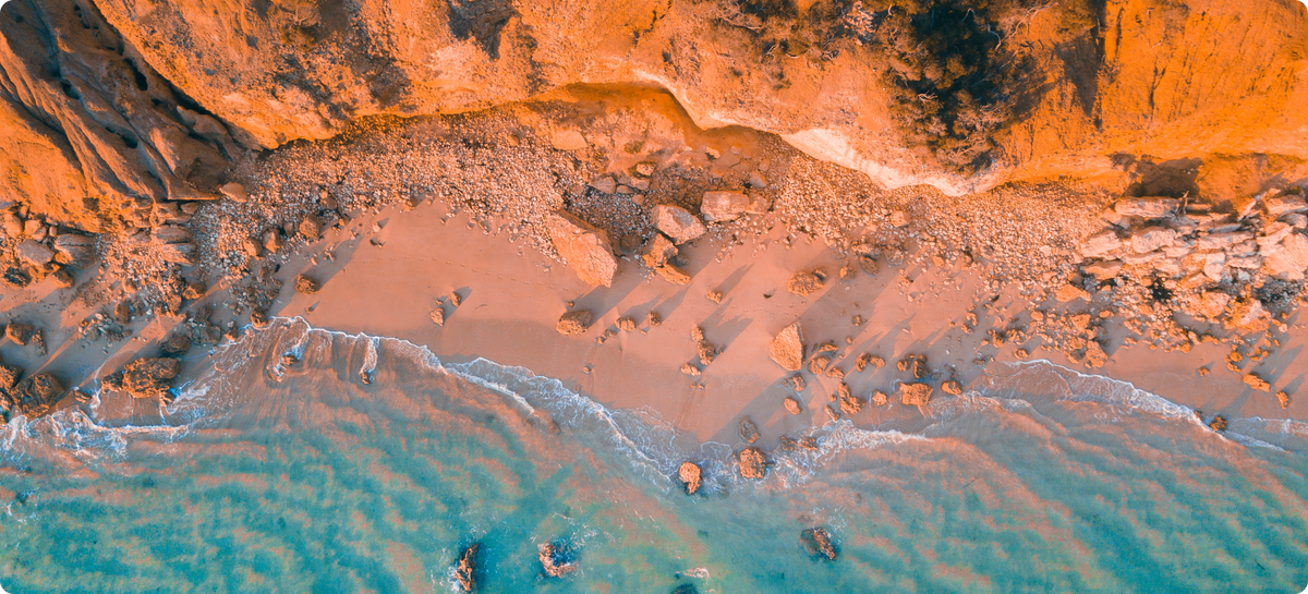 Aerial view of a sandy beach with light brown sand, dark orange cliffs, and turquoise water gently lapping against rocky shorelines.